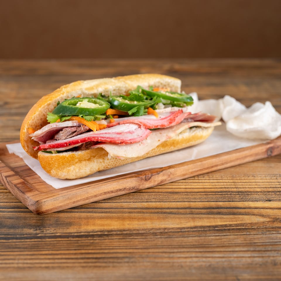 A Vietnamese banh mi sandwich with various meats, cilantro, carrots, and jalapeños on a wooden board.