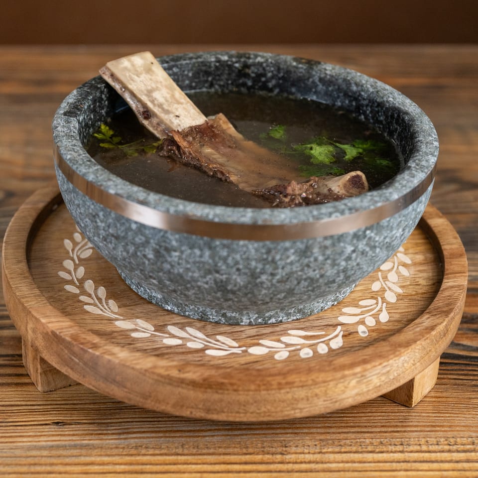 A stone bowl of Vietnamese pho with a large bone and green herbs on a wooden tray.