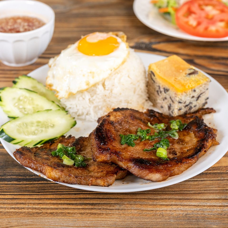 Vietnamese grilled pork chops with rice, a fried egg, cucumber slices, and a savory meatloaf.